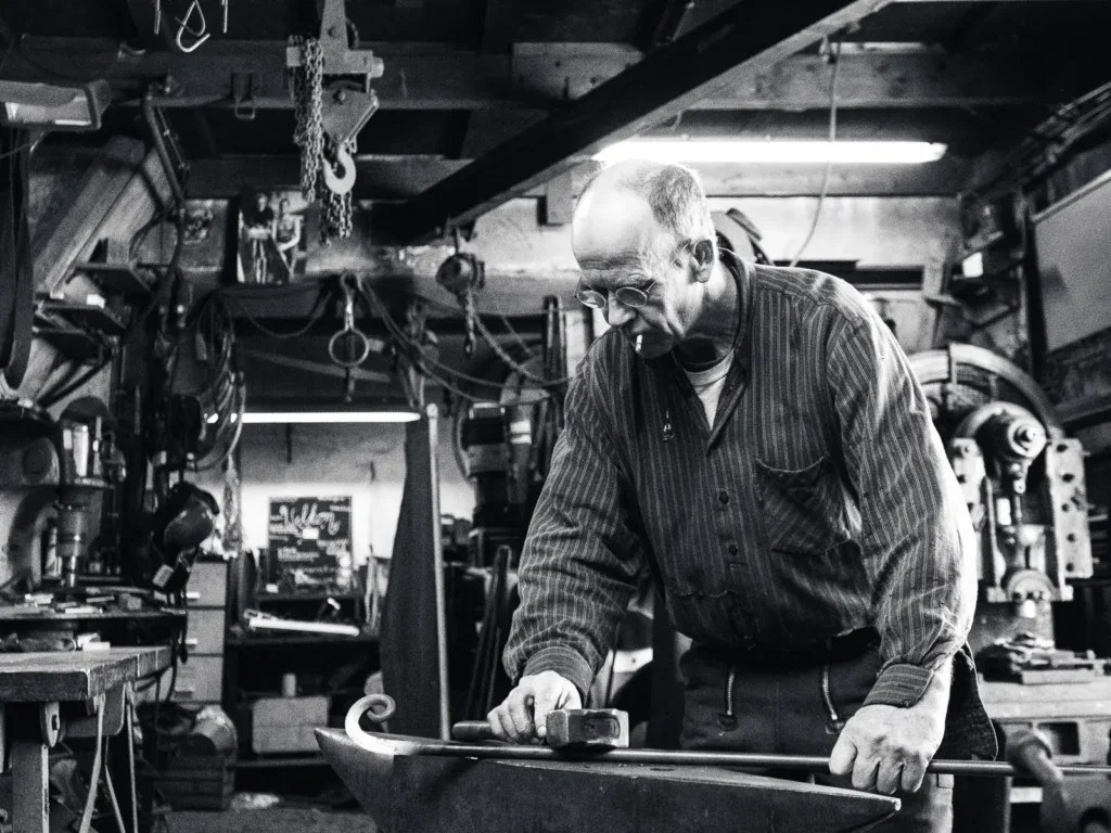Zwart-witfoto van een oudere smid aan het werk in zijn werkplaats, gefotografeerd door Dies